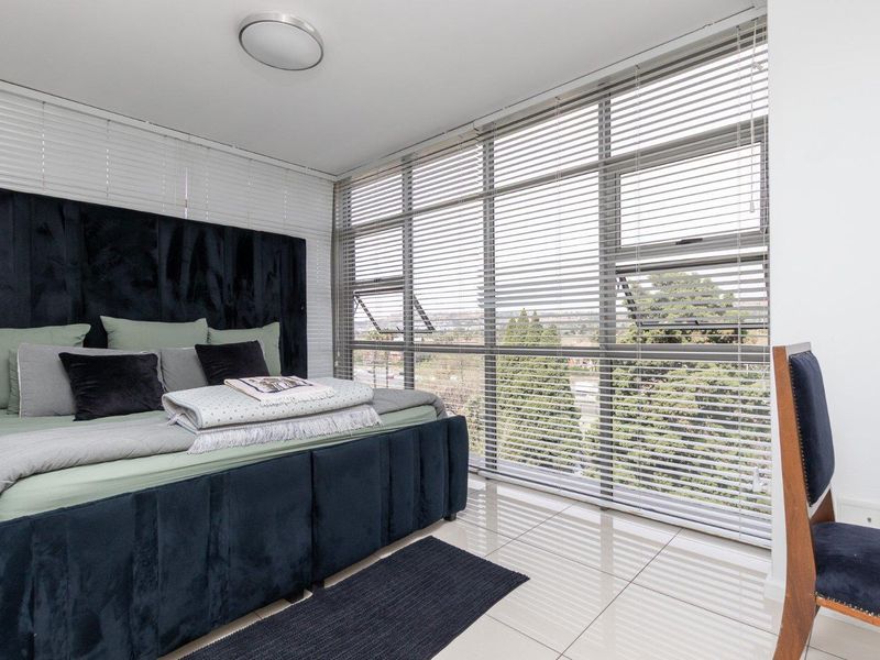A bright bedroom featuring large floor-to-ceiling windows with venetian blinds, polished tiled flooring, and a modern flush mount ceiling light.