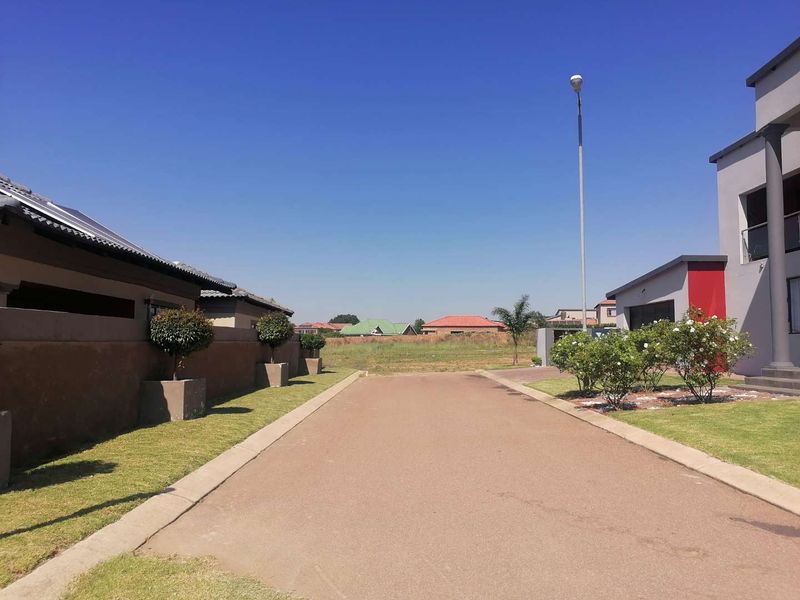 A clear street view showing a paved driveway leading towards a vacant residential plot, flanked by established boundary walls and landscaped garden areas.