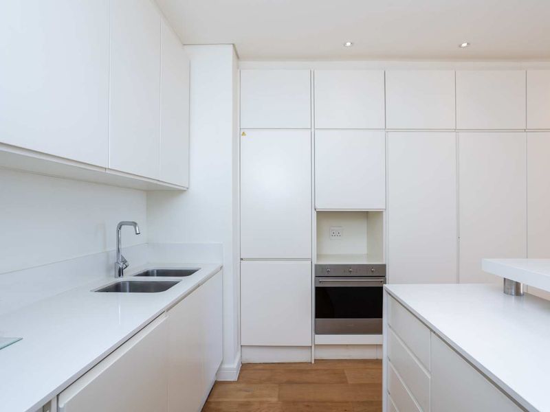 Modern minimalist kitchen featuring sleek white cabinetry, integrated oven, stainless steel sink, and light wood-look flooring with recessed lighting.