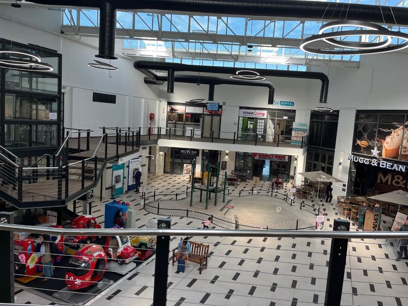 A high-ceilinged, open-plan retail atrium featuring a skylight, exposed industrial ductwork, and multiple commercial storefronts accessible via a central concourse and staircase.