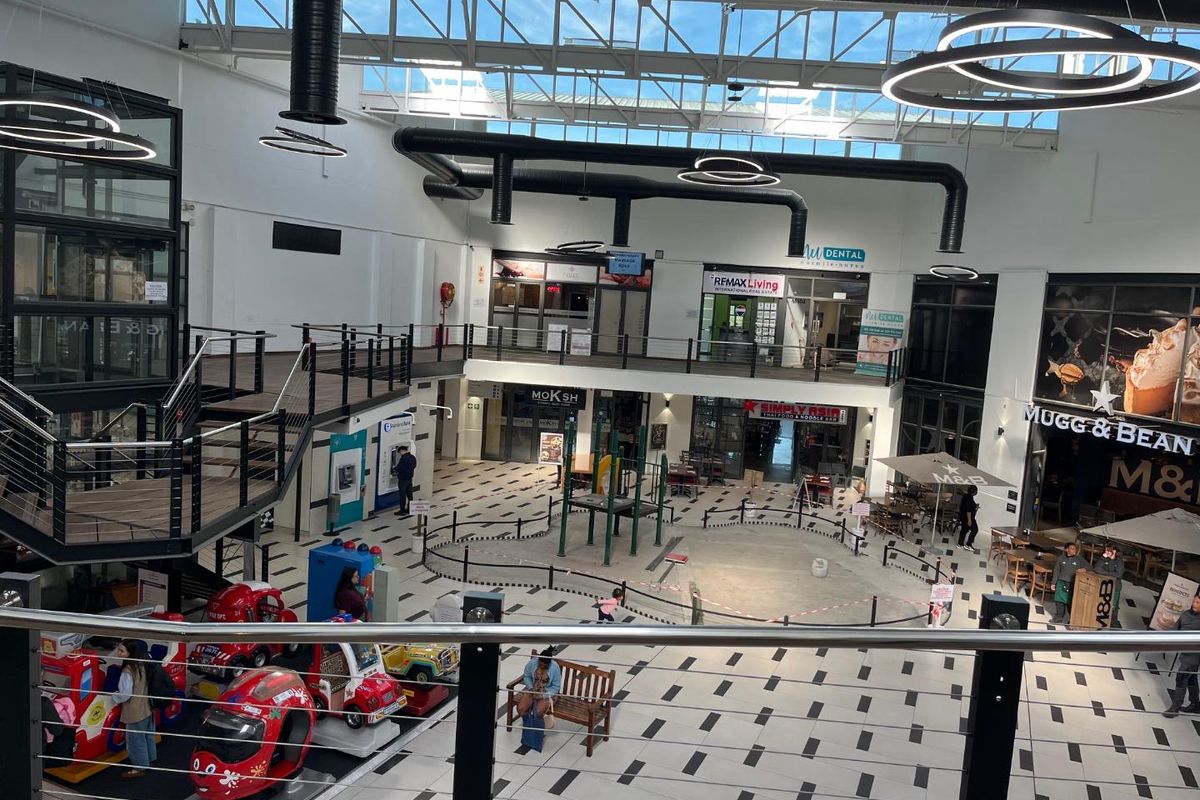 A high-ceilinged, open-plan retail atrium featuring a skylight, exposed industrial ductwork, and multiple commercial storefronts accessible via a central concourse and staircase.