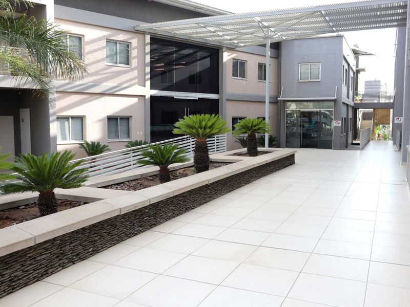 A modern office building exterior featuring a clean tiled walkway, stone-clad planter boxes, and a metal pergola structure leading to a glass entrance.