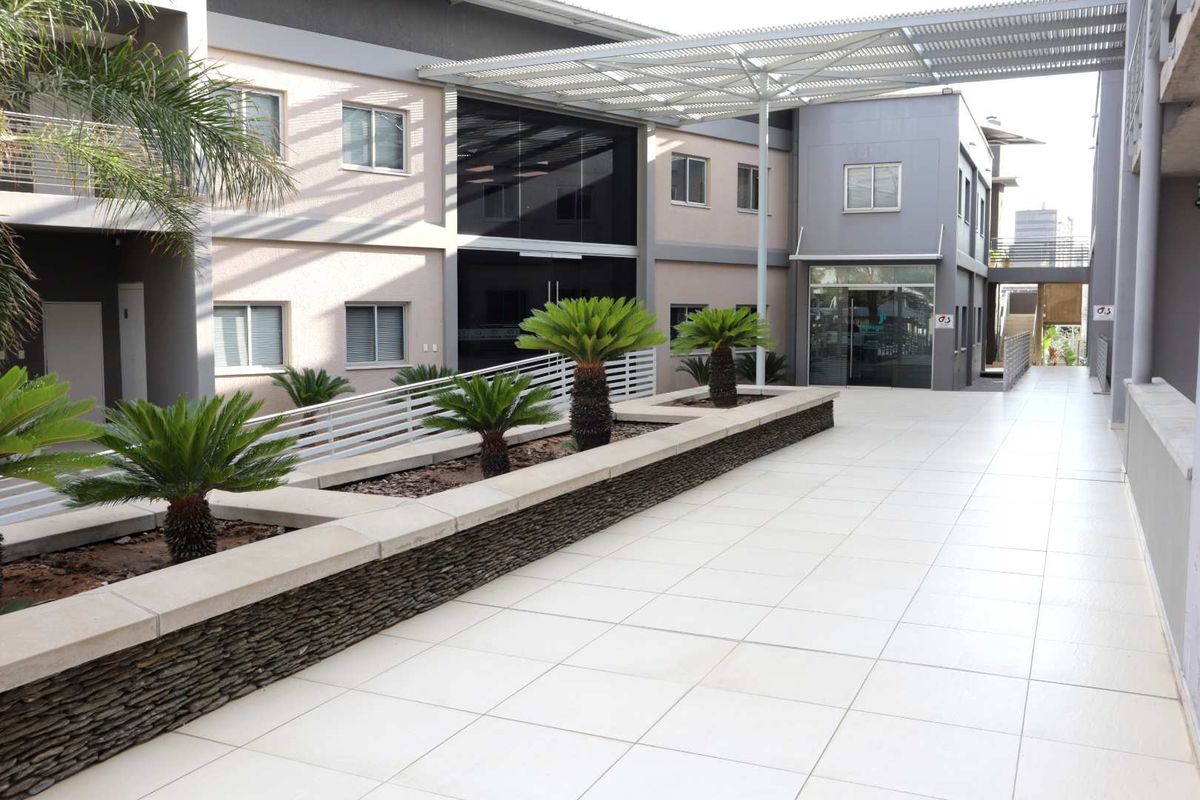 A modern office building exterior featuring a clean tiled walkway, stone-clad planter boxes, and a metal pergola structure leading to a glass entrance.