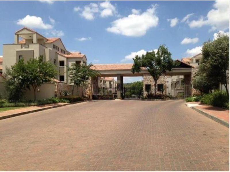 The entrance to the apartment complex features a secure gated access point with stone-clad pillars and a paved driveway.