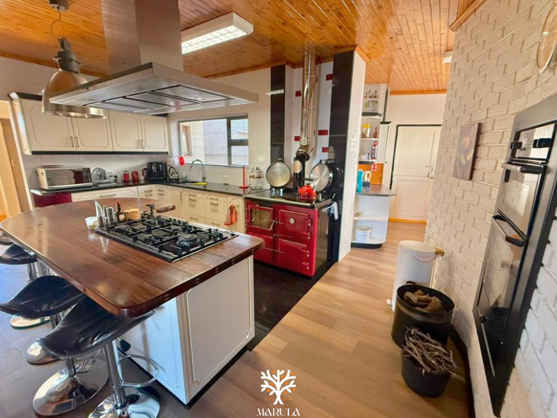 Spacious kitchen featuring a large island with gas hob, integrated oven, wood-burning stove, timber ceiling, and exposed brick accents.