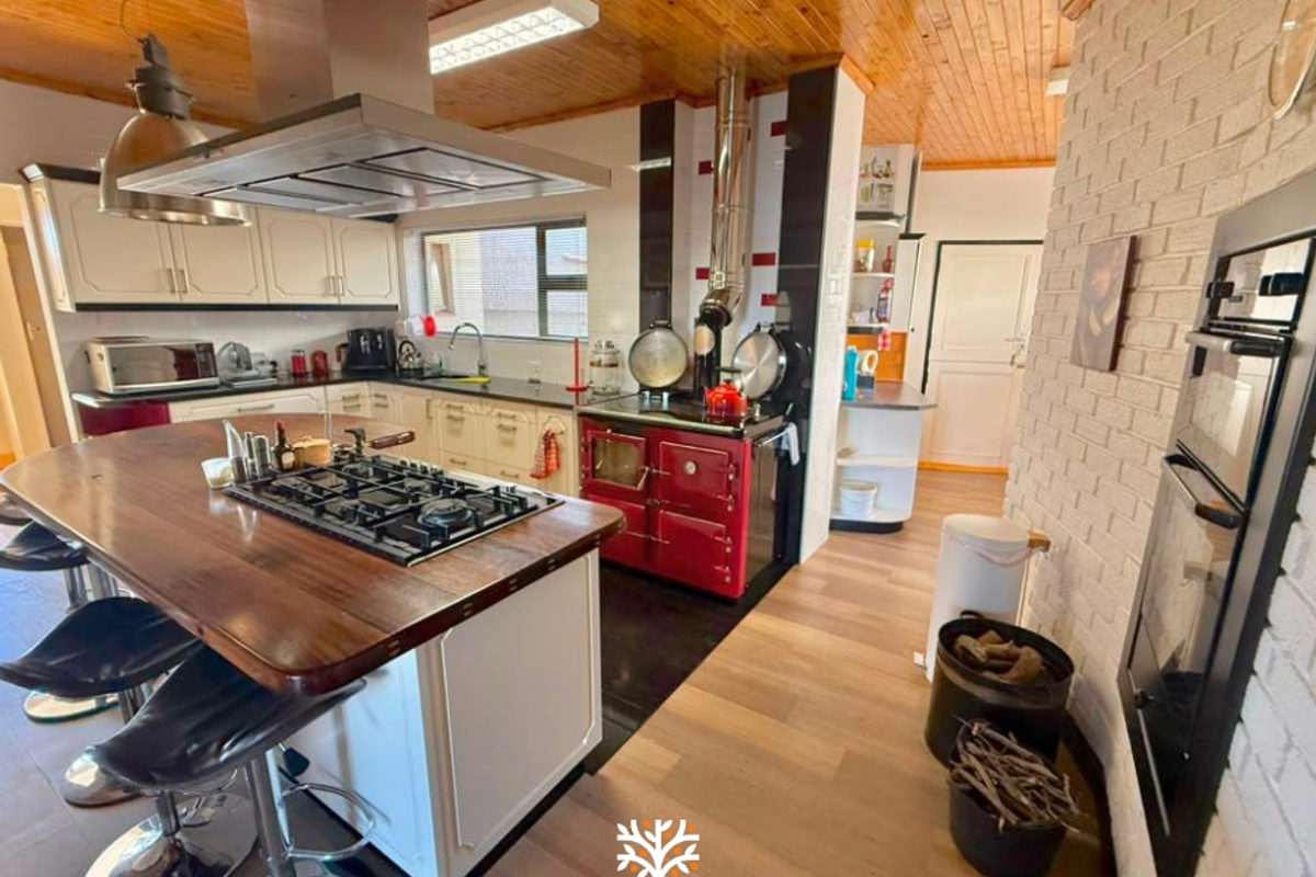 Spacious kitchen featuring a large island with gas hob, integrated oven, wood-burning stove, timber ceiling, and exposed brick accents.