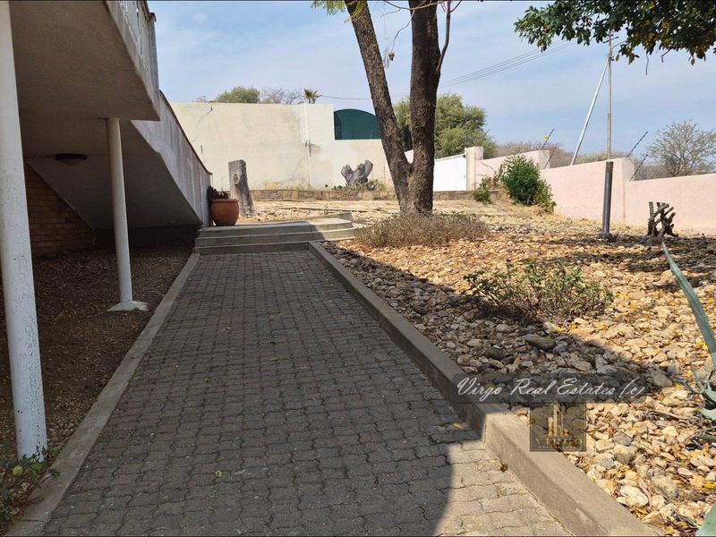 A rear exterior view featuring a paved walkway leading alongside the house, with concrete support columns and a surrounding boundary wall.