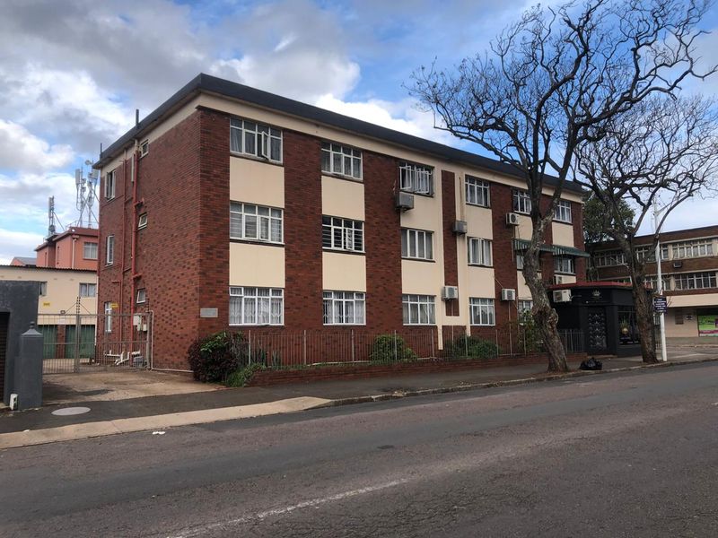 A multi-story brick apartment building featuring security fencing, window bars, and wall-mounted air conditioning units, viewed from the street.