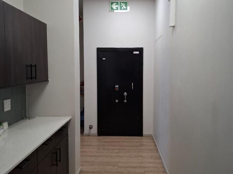 A clean, modern office kitchenette featuring dark wood cabinetry, light countertops, laminate flooring, and a secure metal exit door.