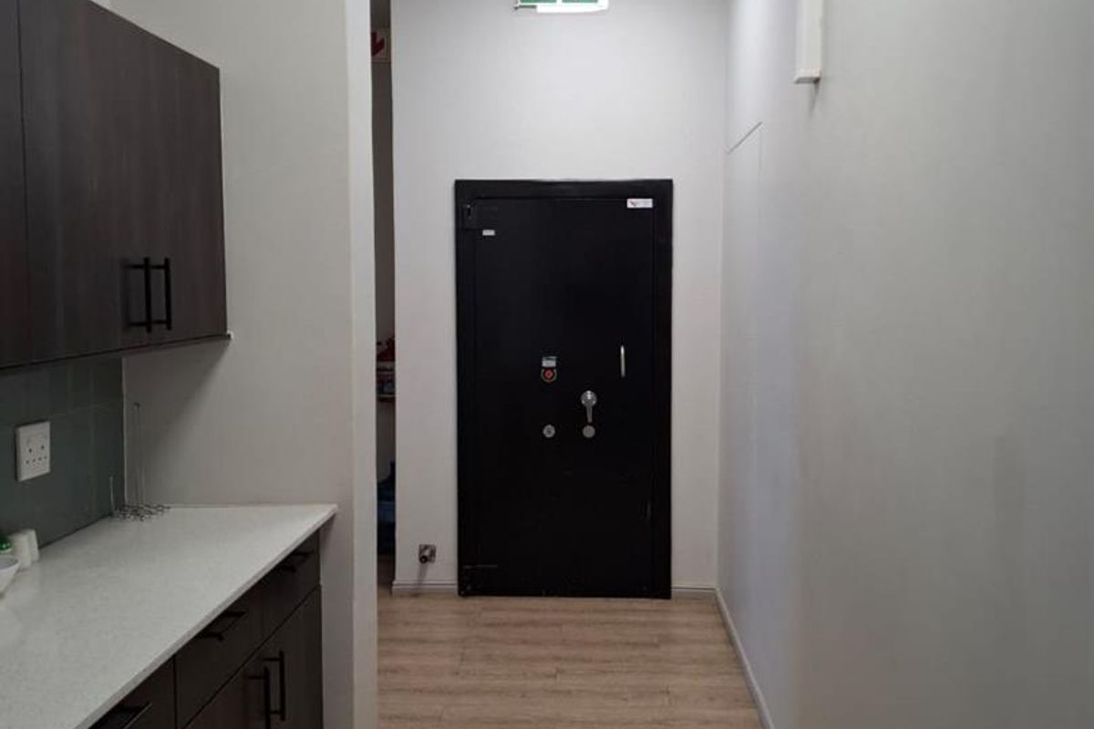 A clean, modern office kitchenette featuring dark wood cabinetry, light countertops, laminate flooring, and a secure metal exit door.
