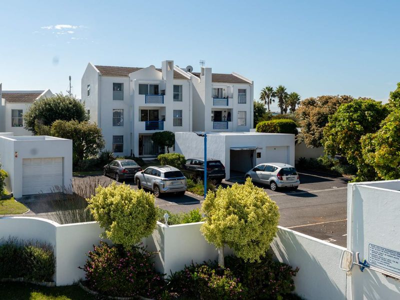 Exterior view of a multi-story apartment complex featuring white facades, private balconies, and an asphalt parking area with individual garage units.