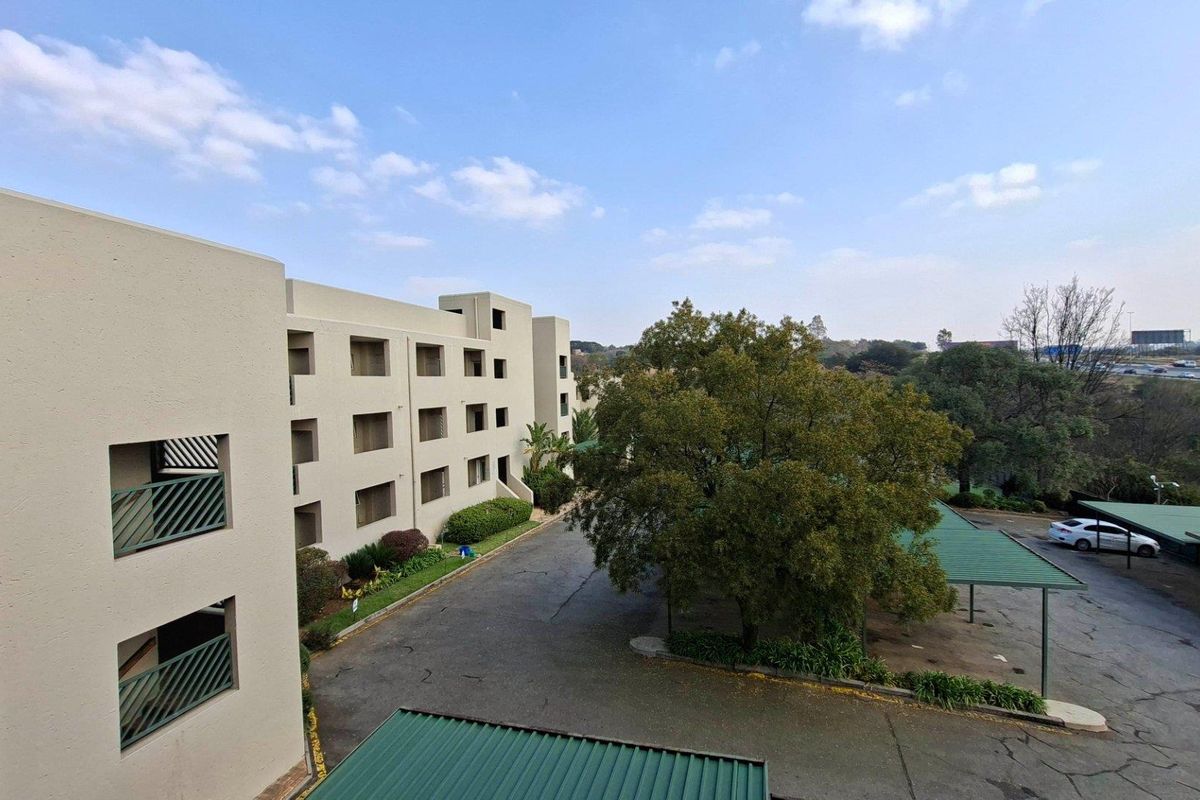 Elevated view of the apartment complex exterior, featuring covered parking bays, paved driveway, and established garden areas.