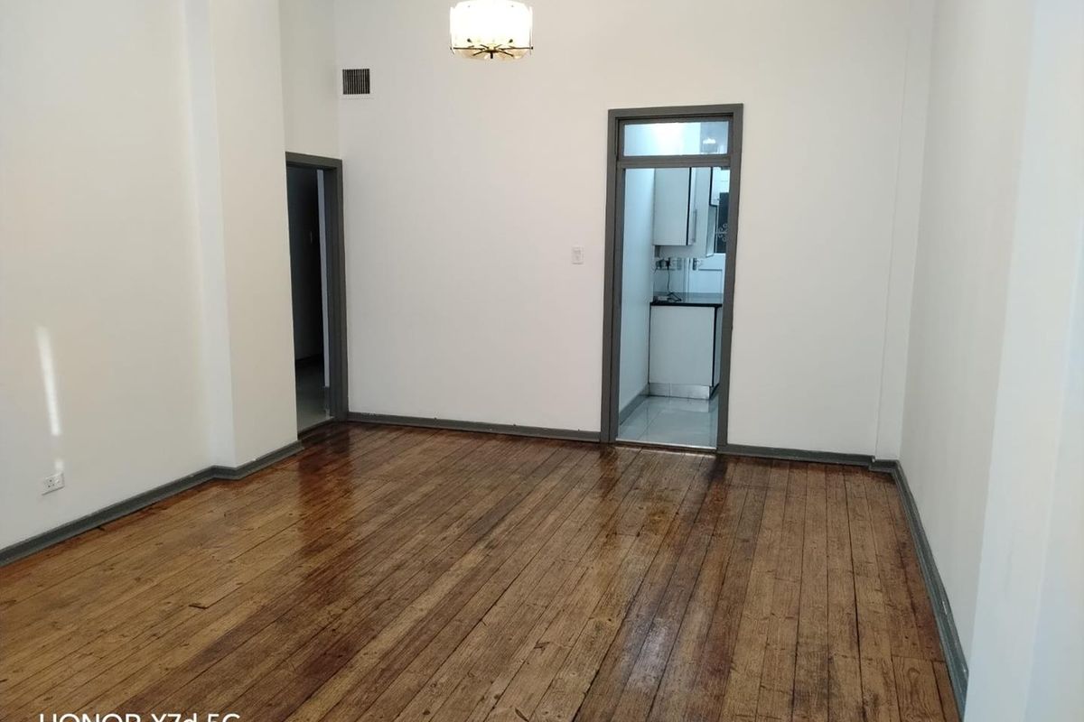 An empty living room featuring polished hardwood flooring, neutral walls, and direct access to an adjacent kitchen area.