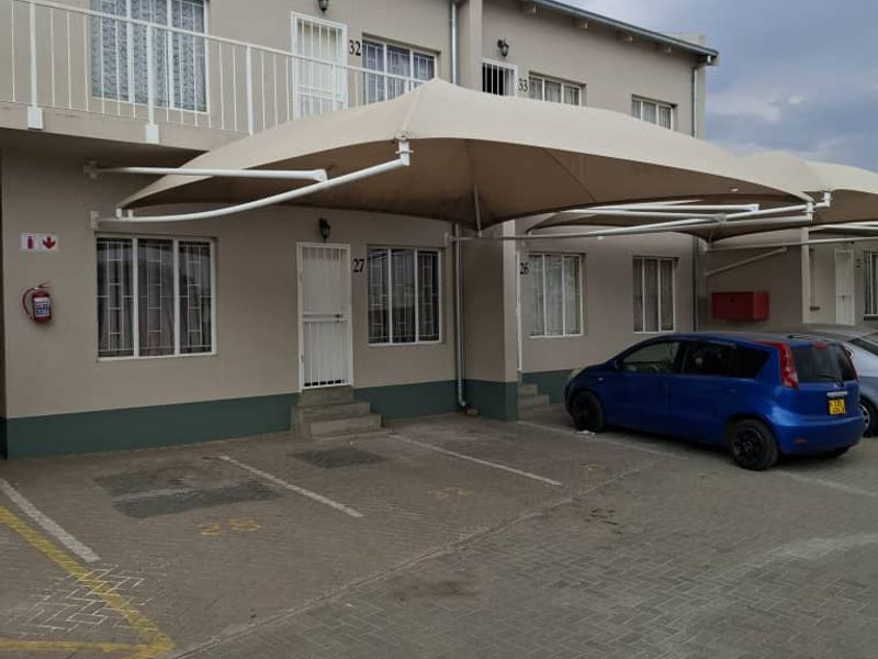 Front exterior view of a multi-story apartment complex featuring paved parking bays, shade sail structures, and security-fitted ground floor entrances.