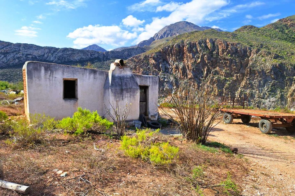 A rustic, weathered farm structure set against a dramatic mountain backdrop, featuring traditional masonry construction and an external chimney.
