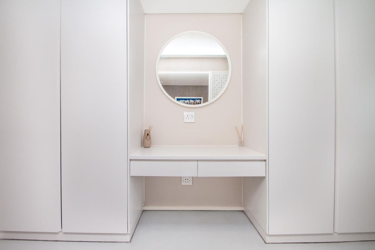 A modern bedroom vanity area featuring built-in floor-to-ceiling cupboards, a floating desk with drawers, and a circular wall-mounted mirror.