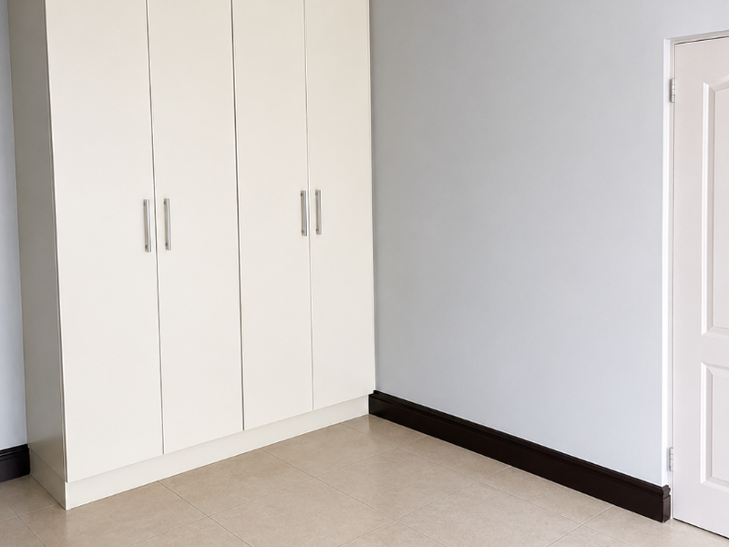 A clean, empty bedroom featuring neutral tiled flooring, built-in cupboards with modern handles, and crisp grey walls with white trim.