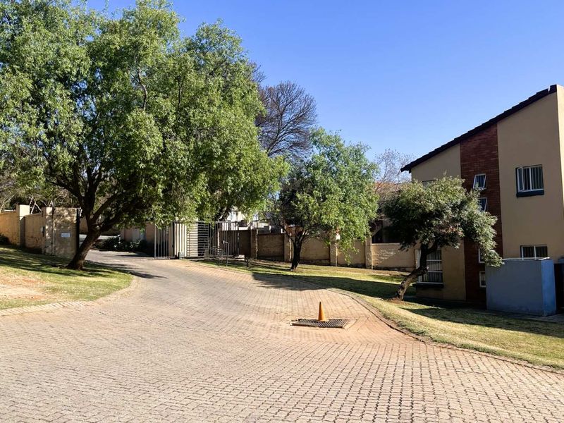 A view of the paved driveway and entrance area of the townhouse complex, featuring established trees and perimeter security walls.
