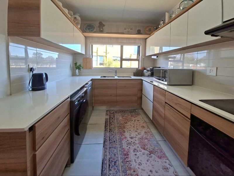 A modern galley-style kitchen featuring wood-grain and white cabinetry, quartz countertops, integrated appliances, and a tiled backsplash with a functional double basin sink.