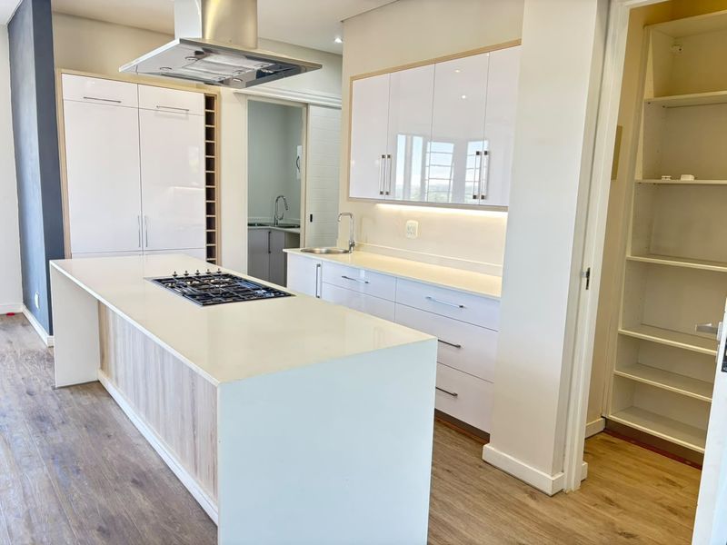 Modern kitchen featuring a central island with gas hob, sleek white cabinetry, stone countertops, and an adjacent walk-in pantry area.