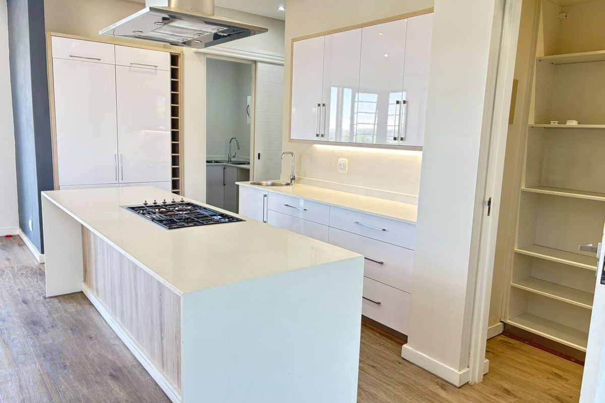 Modern kitchen featuring a central island with gas hob, sleek white cabinetry, stone countertops, and an adjacent walk-in pantry area.