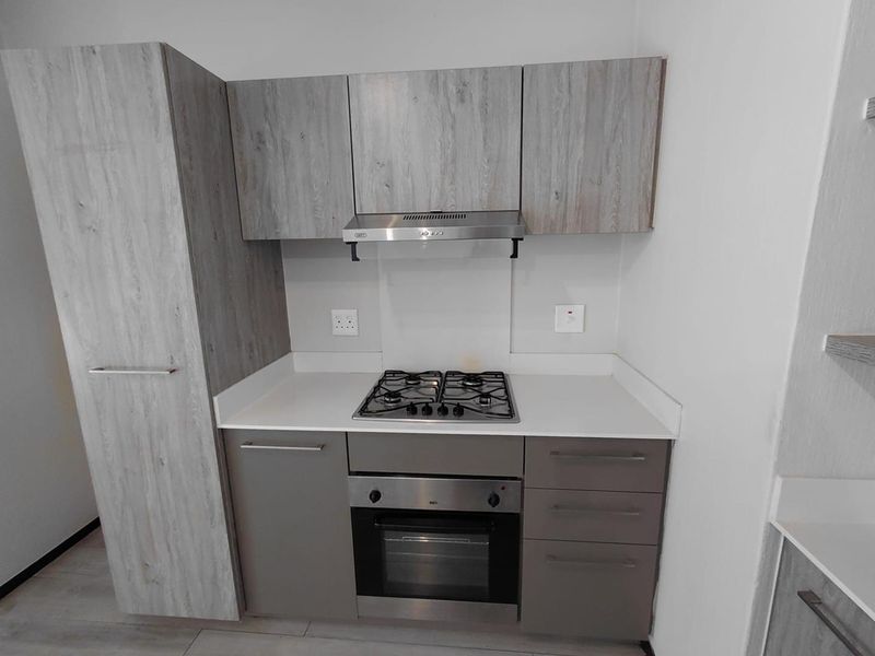 A compact, modern kitchen featuring light wood-grain cabinetry, stone countertops, a built-in gas hob, oven, and stainless steel extractor fan.