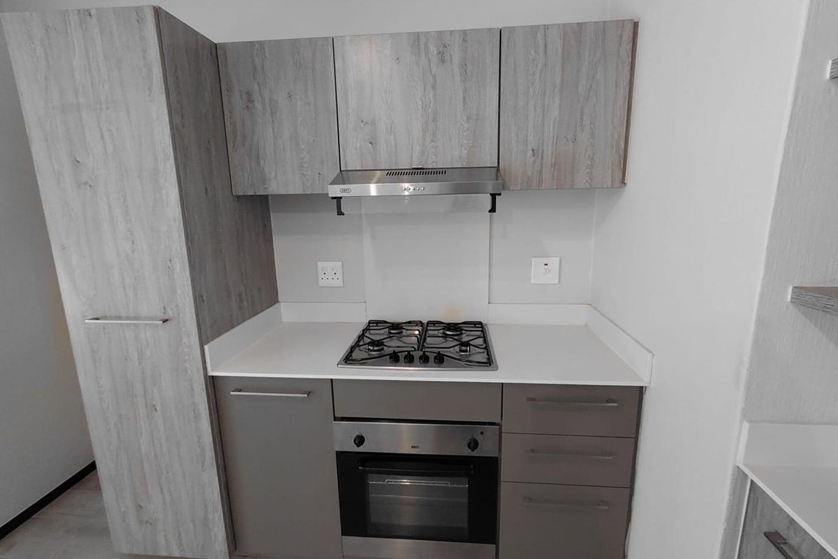 A compact, modern kitchen featuring light wood-grain cabinetry, stone countertops, a built-in gas hob, oven, and stainless steel extractor fan.