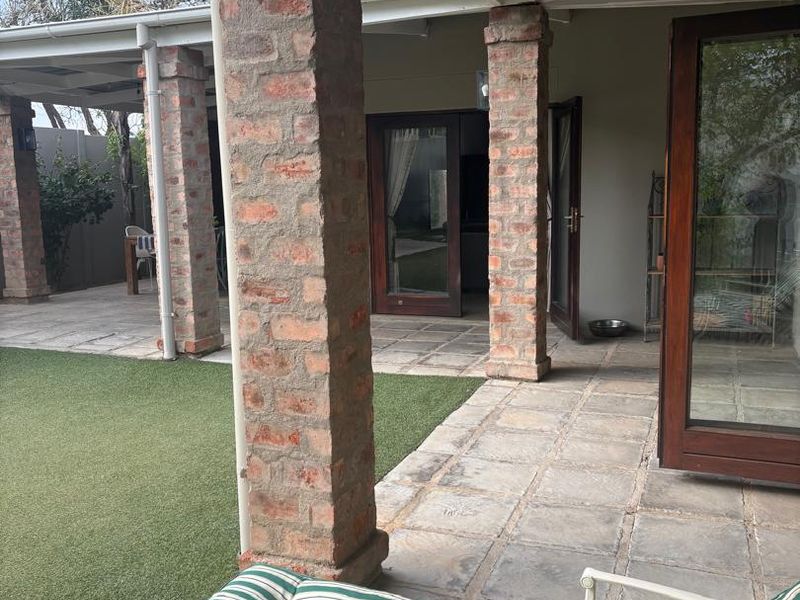 A covered patio area featuring exposed brick columns, stone paving, and timber-framed glass doors providing access to the interior living space.
