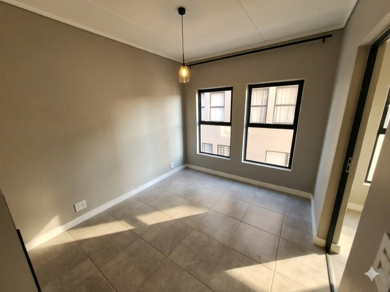 A bright, empty bedroom featuring neutral wall tones, large windows with black frames, and tiled flooring.