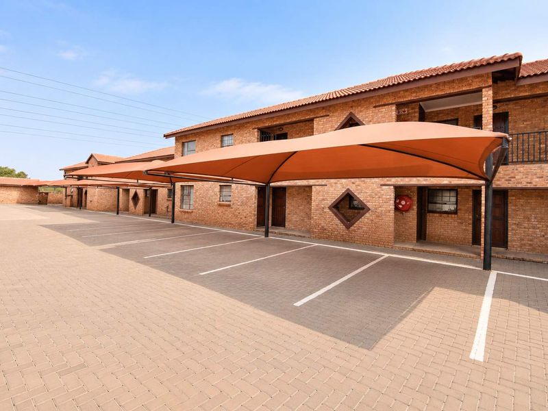Exterior view of an apartment complex featuring a brick facade, tiled roof, and dedicated paved parking bays with shade cloth carports.