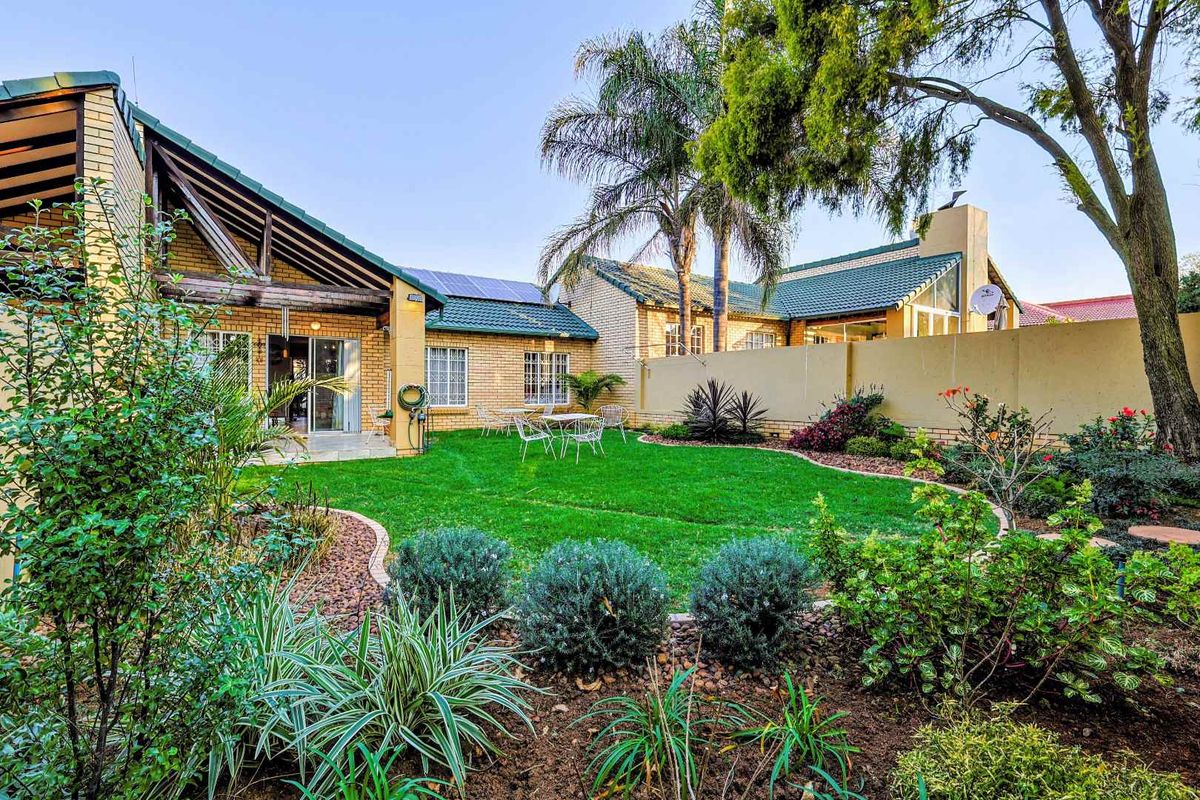 A well-maintained rear garden featuring a lush lawn, landscaped garden beds, a paved patio area, and a brick townhouse facade with solar panels.