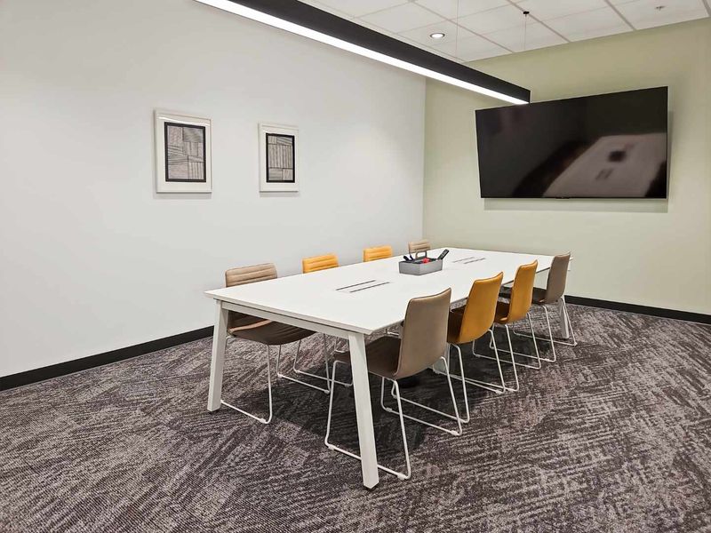 A modern, well-maintained office meeting room featuring a suspended ceiling, linear pendant lighting, carpeted flooring, and a wall-mounted television.