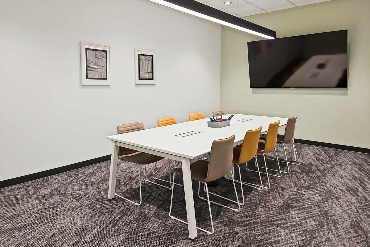 A modern, well-maintained office meeting room featuring a suspended ceiling, linear pendant lighting, carpeted flooring, and a wall-mounted television.