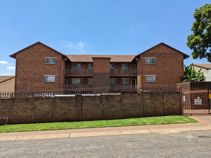 Front exterior view of a multi-story brick apartment complex featuring balconies, secure perimeter walling with razor wire, and gated access.