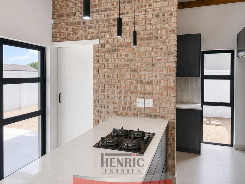 Modern kitchen featuring an exposed brick wall, stone countertops, integrated gas hob, and sleek dark cabinetry with large windows providing natural light.