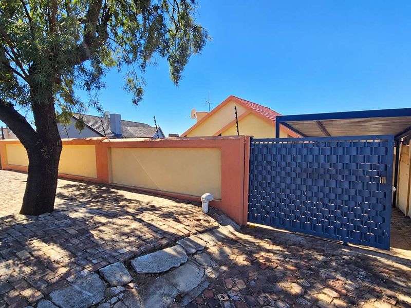 Front exterior view featuring a paved driveway, secure metal gate, carport, and perimeter wall with electric fencing.