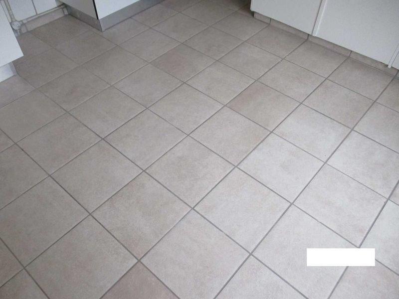 A close-up view of neutral-toned ceramic floor tiles in a kitchen area with white base cabinetry.
