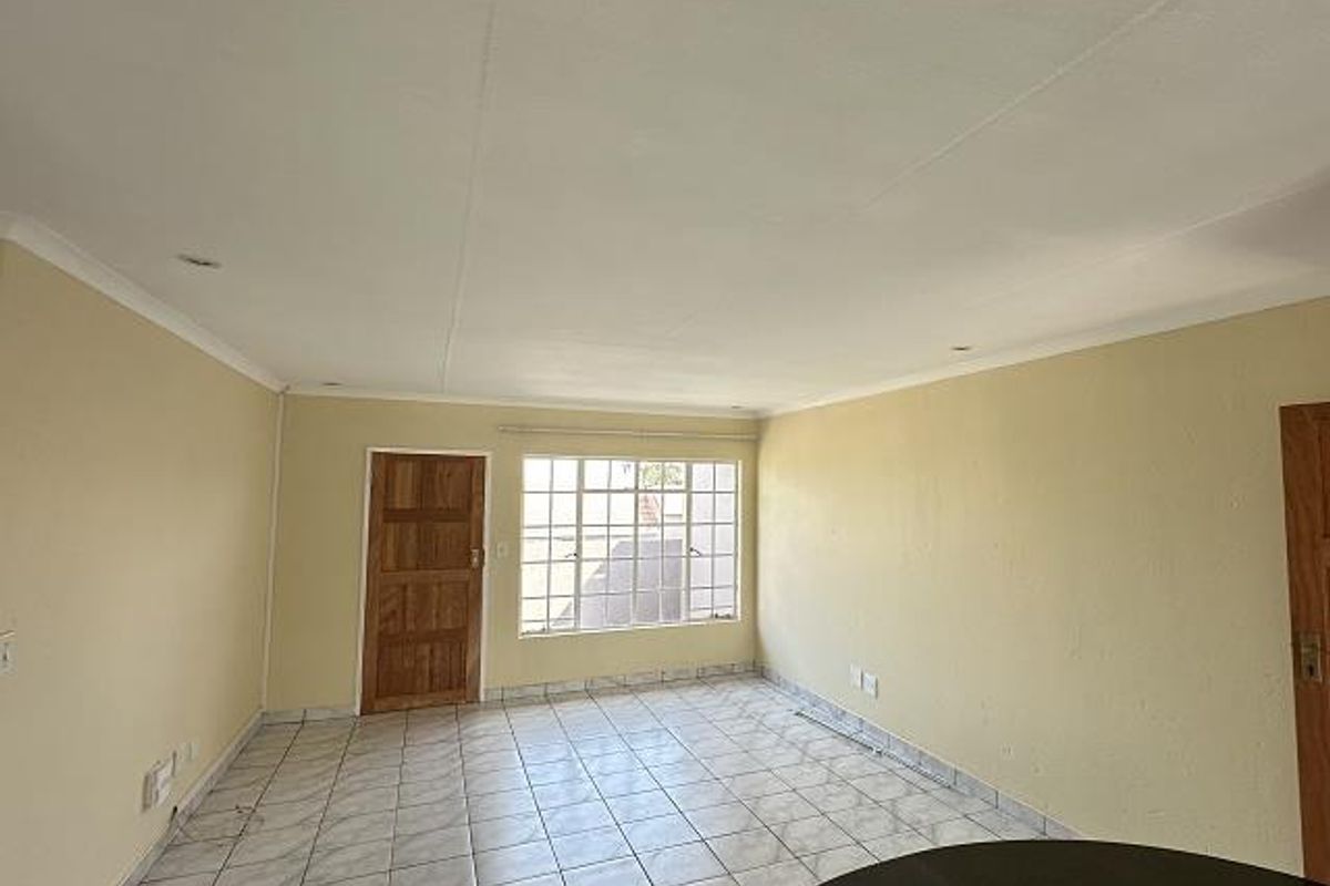 A spacious, empty living area featuring tiled flooring, a wooden door, and large windows that provide natural light.