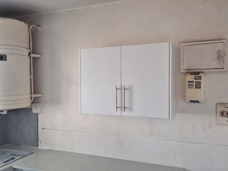 A utility kitchen area featuring a wall-mounted geyser, storage cabinetry, and electrical infrastructure, requiring cosmetic updates to improve the overall finish.