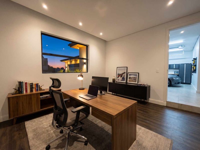 A modern, well-lit home office featuring dark wood-look flooring, recessed lighting, and a view through the window, with direct access to the garage.