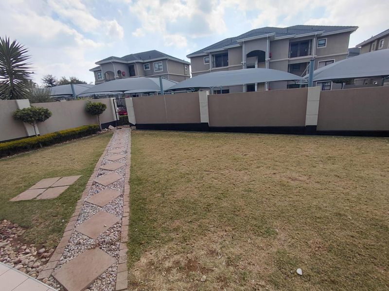A well-maintained rear garden area featuring a paved walkway, perimeter walling, and covered parking structures for the apartment complex.