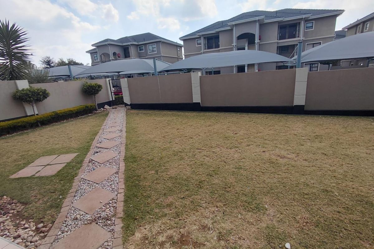 A well-maintained rear garden area featuring a paved walkway, perimeter walling, and covered parking structures for the apartment complex.