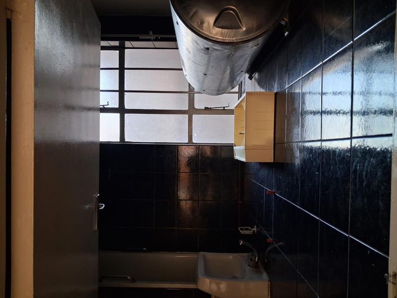 A dated bathroom featuring dark wall tiles, a bathtub, a washbasin, and an exposed wall-mounted water heater.