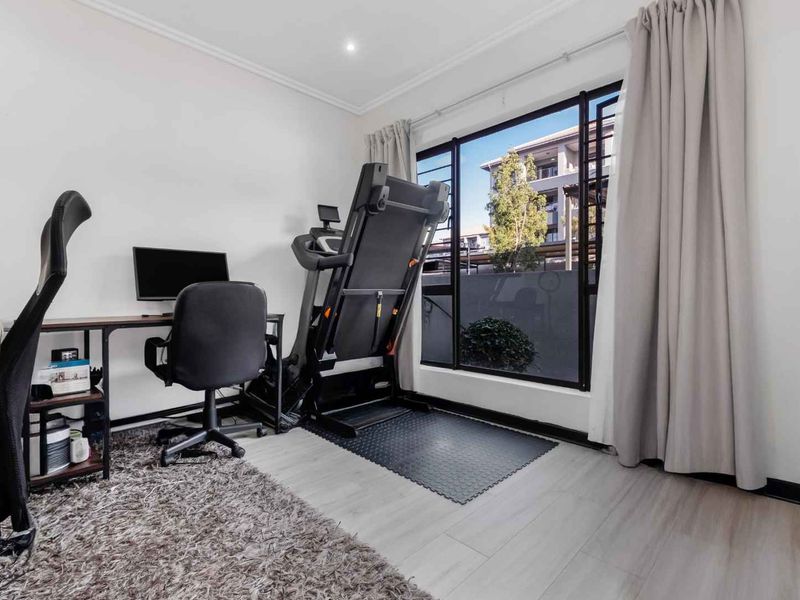 A bright bedroom featuring large windows, modern laminate flooring, and recessed ceiling lighting, currently utilized as a multi-purpose home office and fitness space.