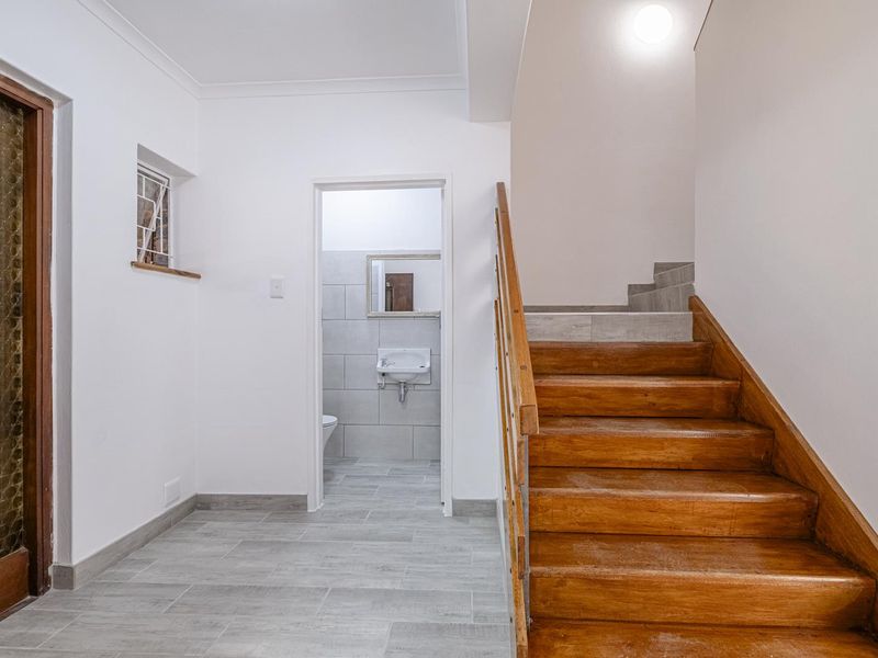 A bright hallway featuring a wooden staircase with a handrail, tiled flooring, and direct access to a small guest bathroom.
