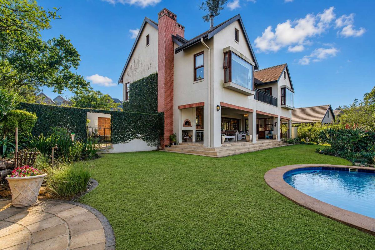 Rear view of a multi-story residence featuring a swimming pool, landscaped garden, covered patio with built-in braai, and a prominent brick chimney.
