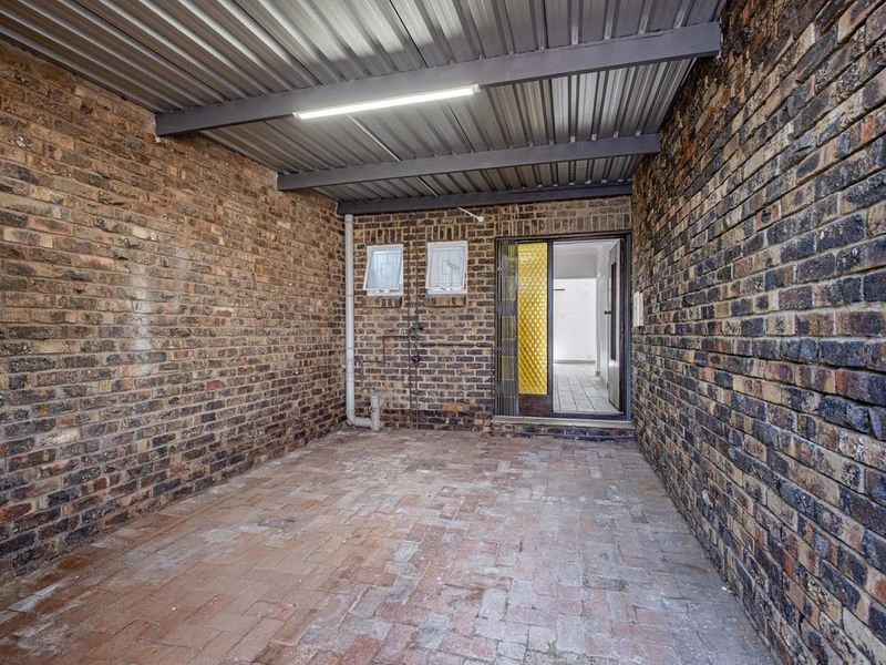 A covered outdoor patio area featuring exposed brick walls, brick paving, and a corrugated metal roof, providing a functional transition space to the interior.