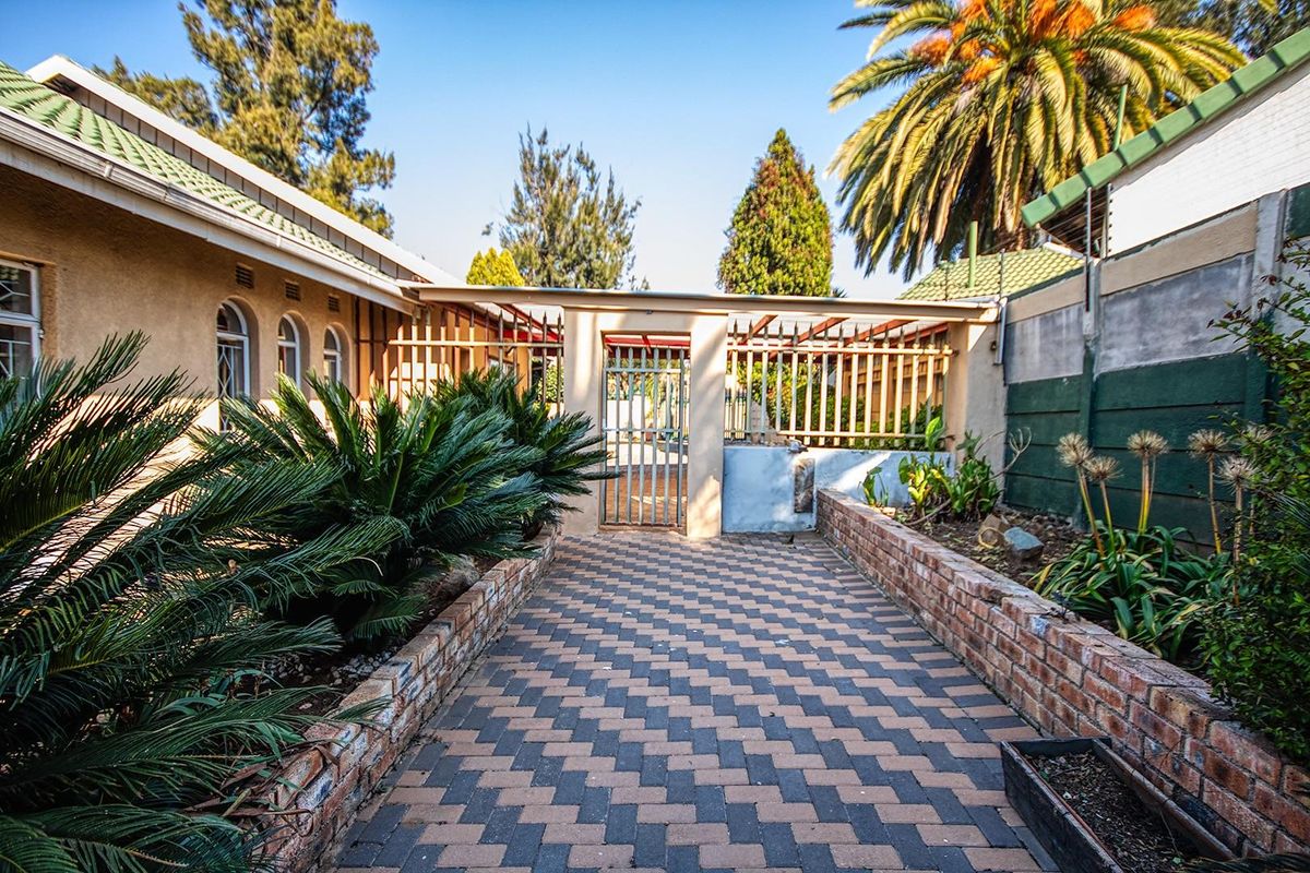 A rear exterior view featuring a brick-paved walkway, landscaped garden beds with retaining walls, and secure metal fencing leading to a gated entrance.