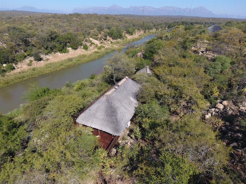 Aerial view of a thatched-roof lodge nestled in a natural bushveld setting alongside a river, offering a serene and secluded environment.
