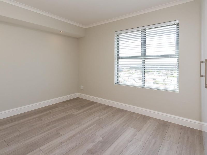 A bright and modern empty bedroom featuring light-toned laminate flooring, neutral walls, and a built-in cupboard with a window providing natural light.